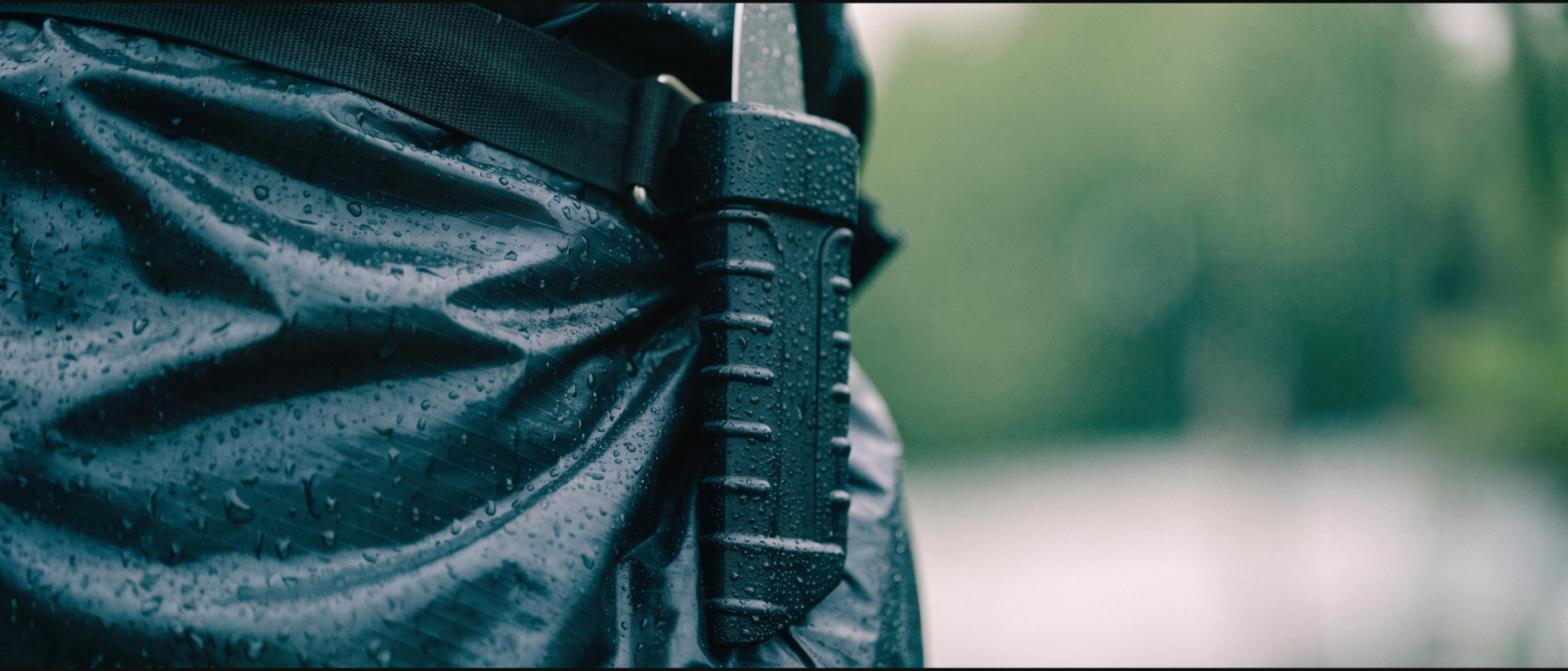A black moulded thermoplastic sheath clipped to a belt over a wet rain jacket, water beaded on the surface